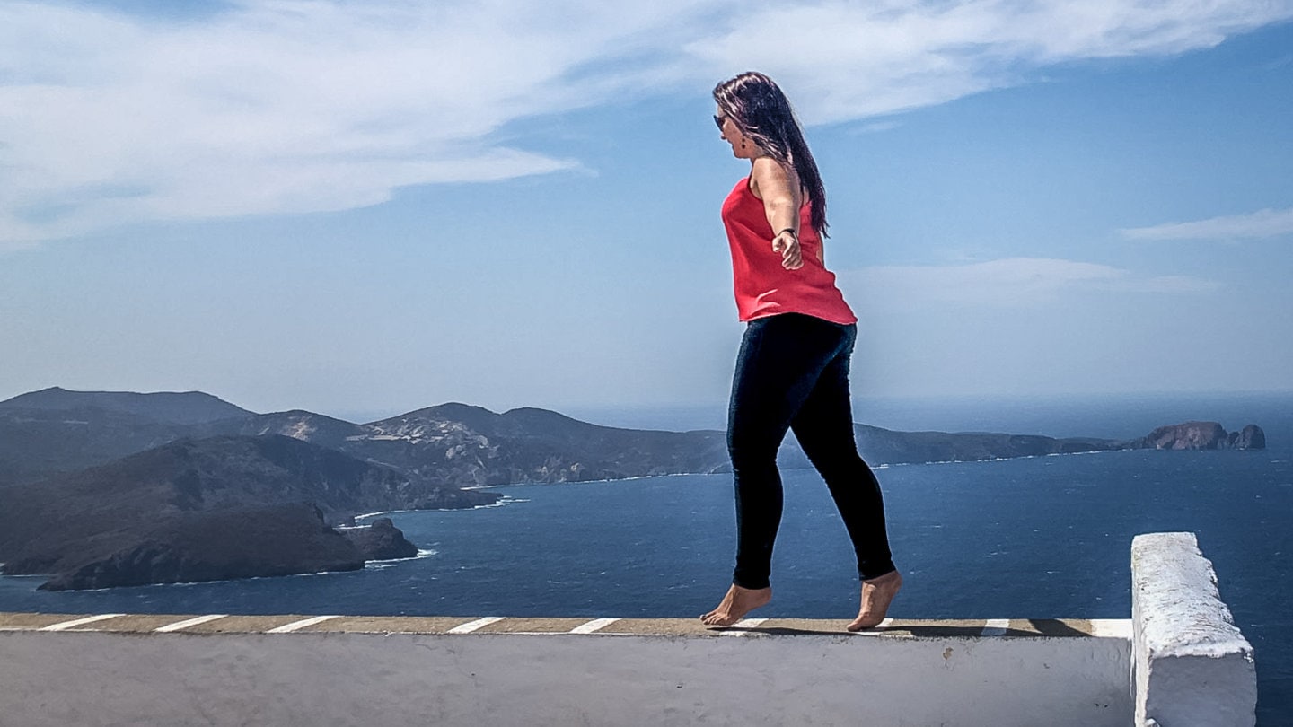 girl walking on a wall with a view of Milos bay behind her